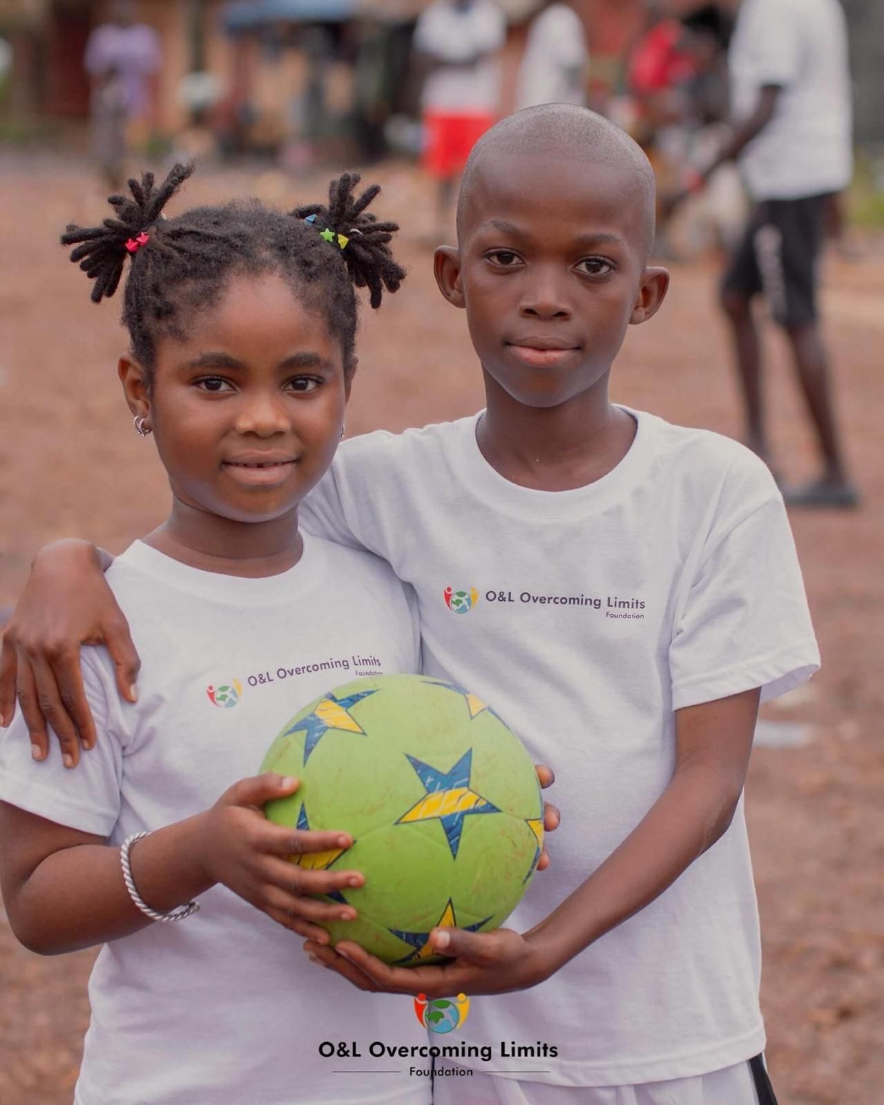 Zwei Kinder in O&L-Overcoming-Limits-Foundation-Shirts halten gemeinsam einen Fußball und blicken lächelnd in die Kamera